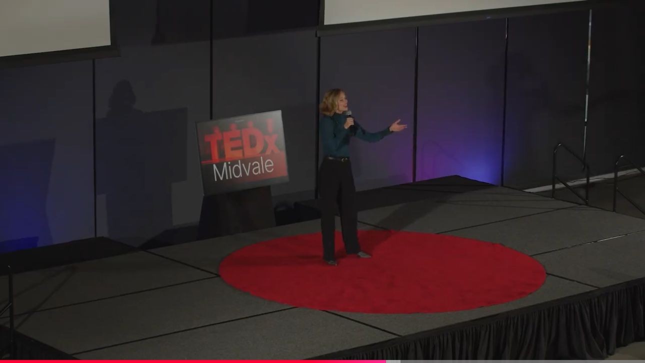 Person speaking on stage at TEDx Midvale event, standing on a red circle carpet.